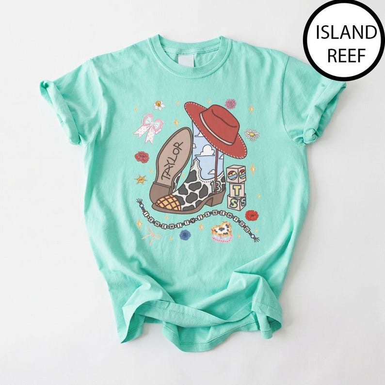 May include: A mint green t-shirt featuring a graphic print of a red cowboy hat, a cow-print cowboy boot with the name Taylor, and a stack of blocks with the letters T, S, and 13. The design includes a beaded string, flowers, and a bow.