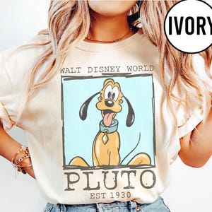 Puede incluir: Sudadera de color marfil con un dise&ntilde;o de Pluto de Walt Disney World. El dise&ntilde;o incluye la imagen de Pluto, las palabras "WALT DISNEY WORLD" y "PLUTO EST 1930" en un marco rectangular. La sudadera tiene mangas largas.