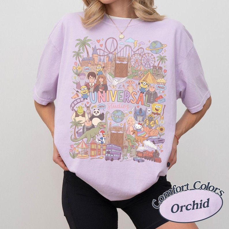 Może przedstawiać: Jasnofioletowy t-shirt z kolorowym nadrukiem graficznym przedstawiającym postacie i punkty orientacyjne Universal Studios. Wz&oacute;r zawiera r&oacute;żne animowane postacie, kolejki g&oacute;rskie oraz napis Universal Studios wielokolorową czcionką.