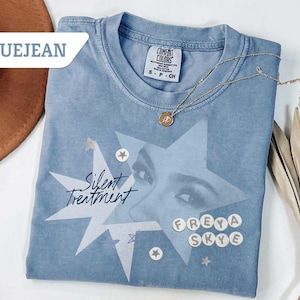May include: Blue jean colored t-shirt with a graphic design featuring a star, eyes, and the words "Silent Treatment" and "Freya Skye." A gold necklace with a pendant is draped on the shirt. The shirt is folded and displayed with a brown hat and dried plant.