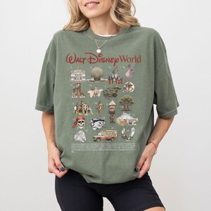 Puede incluir: Una camiseta verde oliva con el texto "Walt Disney World" en rojo. La camiseta muestra ilustraciones de atracciones de Disney World, como un carrusel, Spaceship Earth y el castillo de Cenicienta.