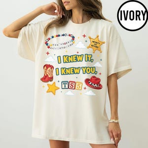 May include: An ivory t-shirt featuring a graphic print with the text I KNEW IT, I KNEW YOU, and TS5. The design includes a beaded bracelet, a gold star badge labeled INFINITY AND BEYOND, a red cowboy boot, a red cowboy hat, and small stars.