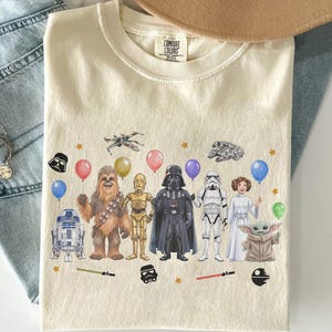 May include: Cream-colored t-shirt featuring a Star Wars-themed graphic. The design includes various characters like Darth Vader, Chewbacca, and R2-D2, along with balloons and stars. The shirt has a Comfort Colors tag.