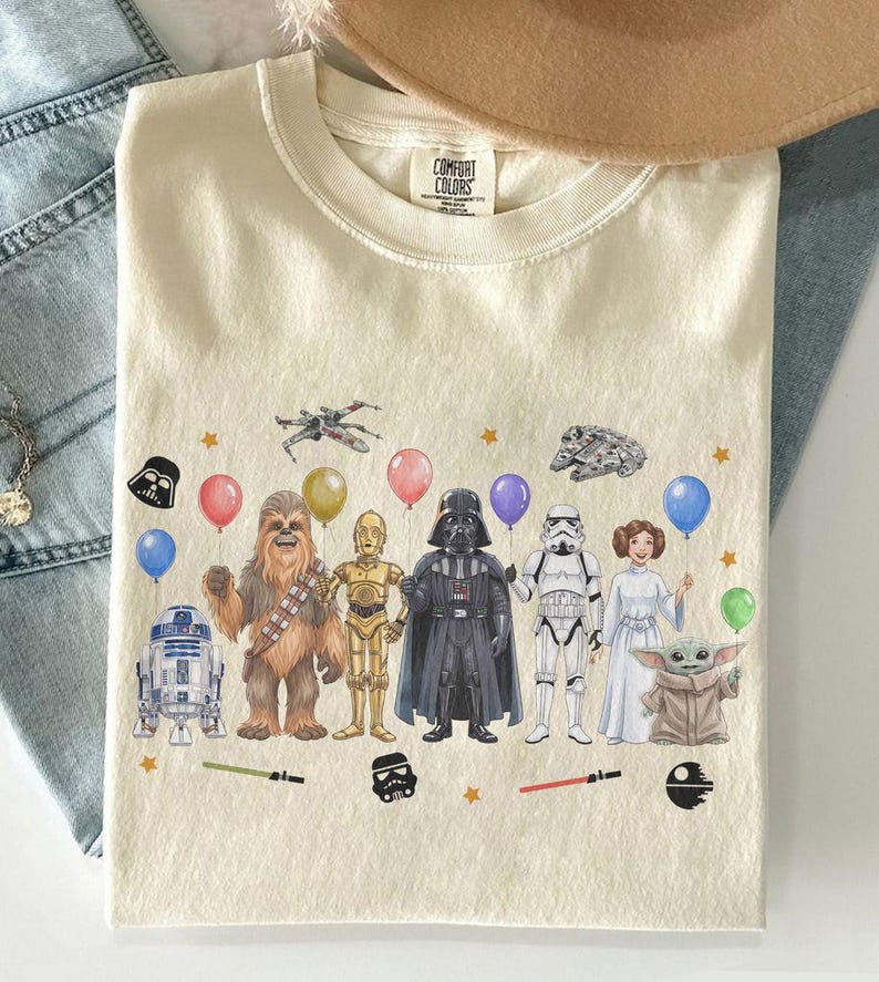 May include: Cream-colored t-shirt featuring a Star Wars-themed graphic. The design includes various characters like Darth Vader, Chewbacca, and R2-D2, along with balloons and stars. The shirt has a Comfort Colors tag.