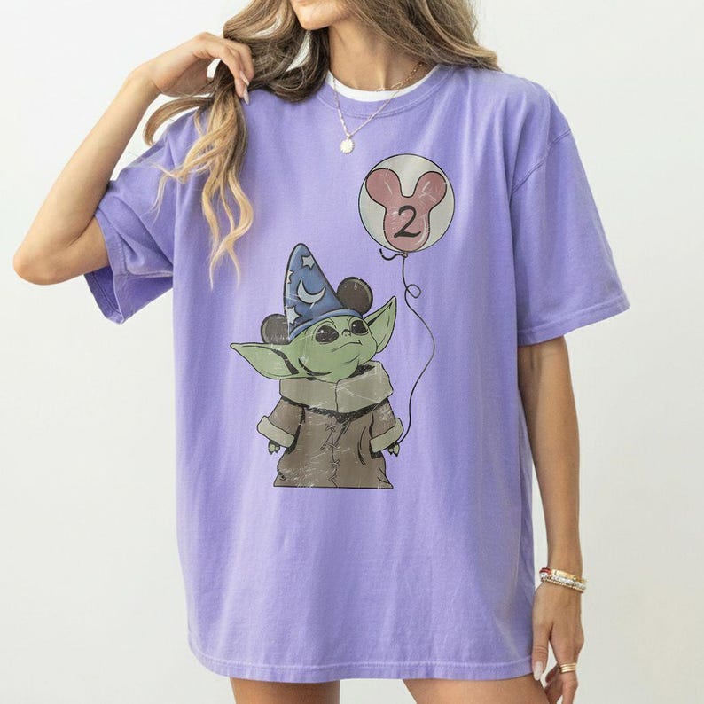 May include: A light purple t-shirt featuring a cartoon character resembling Baby Yoda, wearing a wizard hat and brown coat. A balloon with the number "2" is floating next to the character. The shirt is a solid color with a relaxed fit.