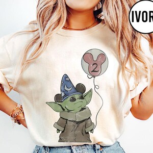 May include: Ivory-colored t-shirt featuring a cartoon Grogu character wearing a wizard hat with Mickey Mouse ears. Grogu is holding a balloon with the number 2. The design is in a vintage style.