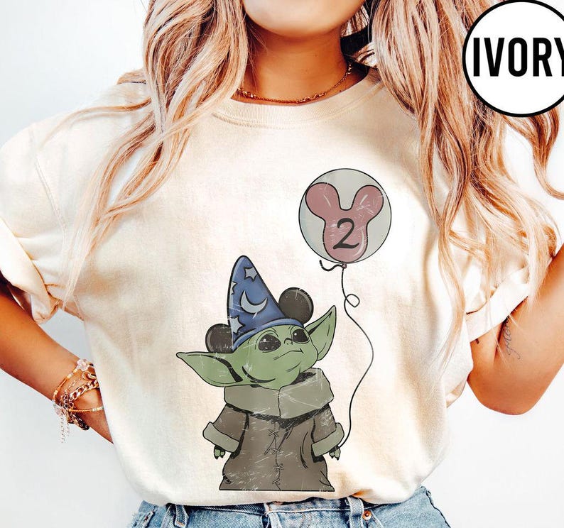 May include: Ivory-colored t-shirt featuring a cartoon Grogu character wearing a wizard hat with Mickey Mouse ears. Grogu is holding a balloon with the number 2. The design is in a vintage style.