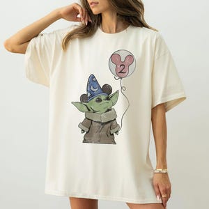 May include: Cream-colored t-shirt featuring a cartoon illustration of a green alien-like character wearing a blue wizard hat with Mickey Mouse ears. The character holds a pink balloon with the number 2. The design is centered on the shirt.