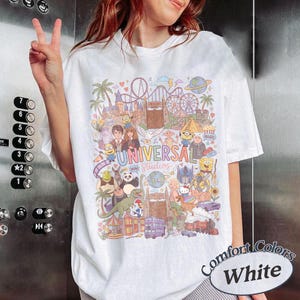 Może przedstawiać: Biały t-shirt z kolorowym nadrukiem graficznym przedstawiającym postacie i zabytki Universal Studios. Wz&oacute;r zawiera ilustracje r&oacute;żnych animowanych postaci, kolejkę g&oacute;rską oraz napis Universal Studios wykonany wielokolorową czcionką na środku.