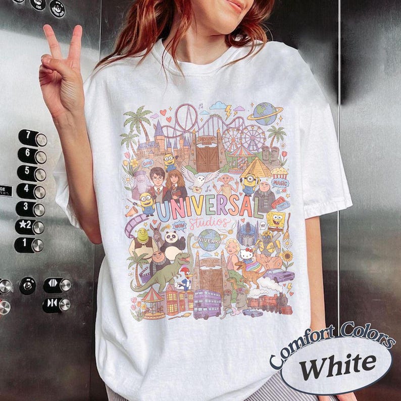 Może przedstawiać: Biały t-shirt z kolorowym nadrukiem graficznym przedstawiającym postacie i zabytki Universal Studios. Wz&oacute;r zawiera ilustracje r&oacute;żnych animowanych postaci, kolejkę g&oacute;rską oraz napis Universal Studios wykonany wielokolorową czcionką na środku.