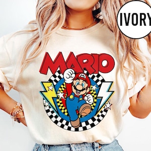 May include: An ivory t-shirt featuring a graphic of the character Mario in a red cap and blue overalls. The design includes the word MARIO in large red letters above a circular checkered pattern with yellow lightning bolts and white stars.