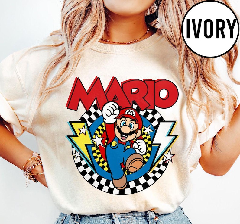 May include: An ivory t-shirt featuring a graphic of the character Mario in a red cap and blue overalls. The design includes the word MARIO in large red letters above a circular checkered pattern with yellow lightning bolts and white stars.