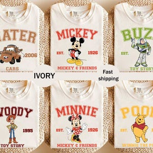 May include: Ivory-colored t-shirts featuring various cartoon characters and text. Designs include "Mater" with a car, "Mickey" with Mickey Mouse, "Buzz" with Buzz Lightyear, "Woody" with Woody, "Minnie" with Minnie Mouse, and "Pooh" with Winnie the Pooh.