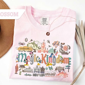Op de afbeelding: Een lichtroze t-shirt met een kleurrijke afbeelding van het Magic Kingdom. De afbeelding bevat stripfiguren, een kasteel en de woorden "Magic Kingdom". Een gouden ketting met een hanger ligt over het shirt.