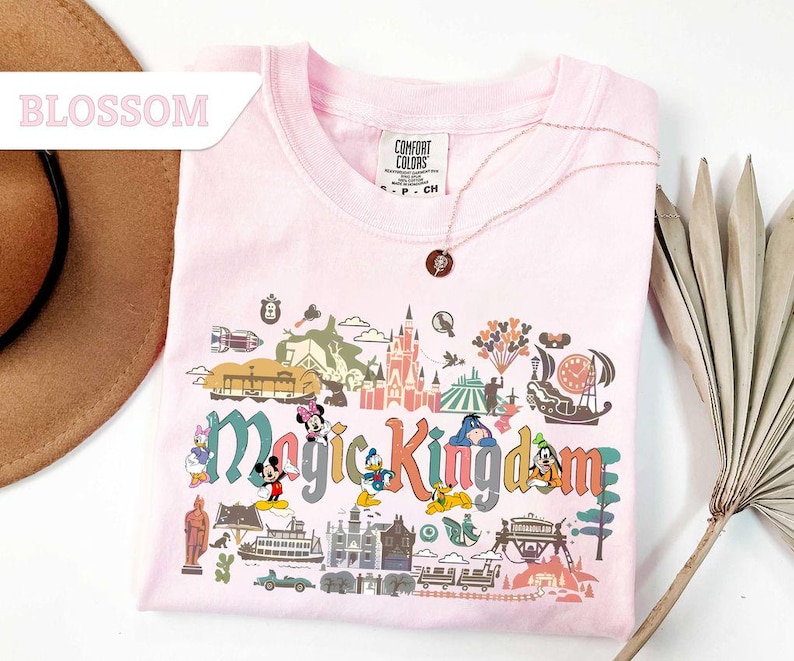 Op de afbeelding: Een lichtroze t-shirt met een kleurrijke afbeelding van het Magic Kingdom. De afbeelding bevat stripfiguren, een kasteel en de woorden "Magic Kingdom". Een gouden ketting met een hanger ligt over het shirt.