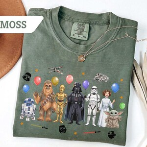 May include: Moss green t-shirt featuring a Star Wars character design. The design includes characters like Darth Vader, Chewbacca, and R2-D2, along with balloons and stars. A gold necklace is draped over the shirt.