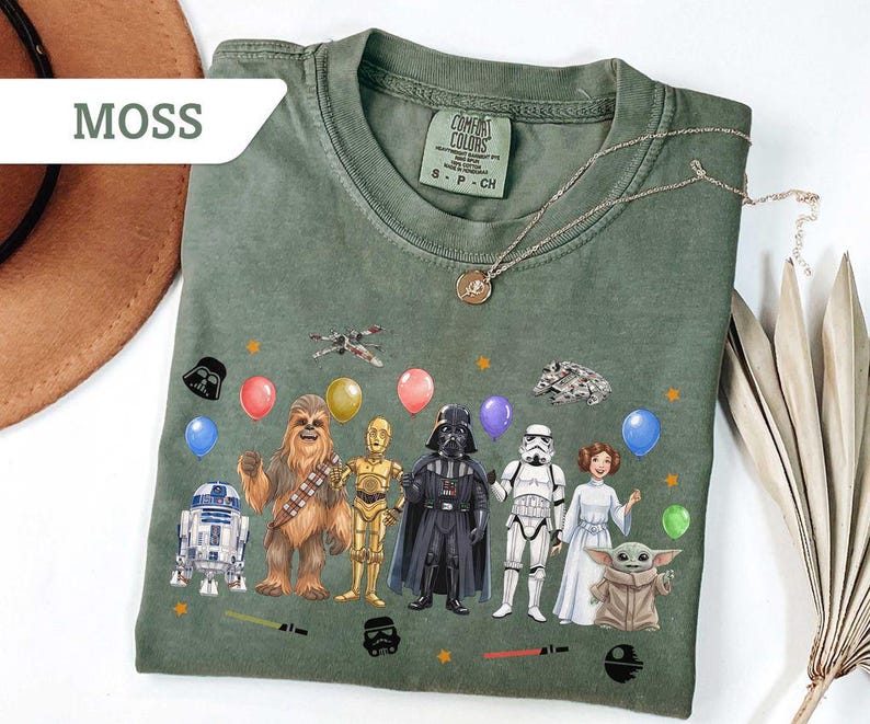May include: Moss green t-shirt featuring a Star Wars character design. The design includes characters like Darth Vader, Chewbacca, and R2-D2, along with balloons and stars. A gold necklace is draped over the shirt.
