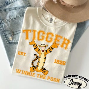 May include: Ivory-colored t-shirt featuring the character Tigger from Winnie the Pooh. The shirt has the word "Tigger" in orange block letters, with "EST. 1926" and "Winnie the Pooh" printed below. The shirt is folded and placed on top of blue jeans.