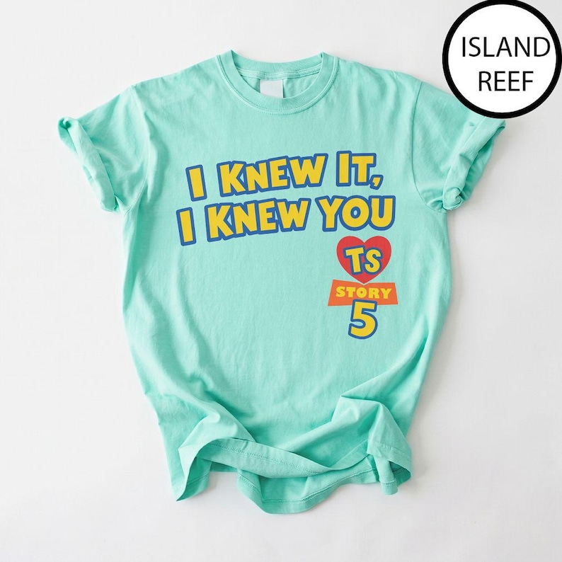 May include: A light mint green short-sleeve t-shirt featuring the yellow text I KNEW IT, I KNEW YOU with a red heart containing the letters TS above the orange text STORY and the large yellow number 5. The shirt has a crew neckline.