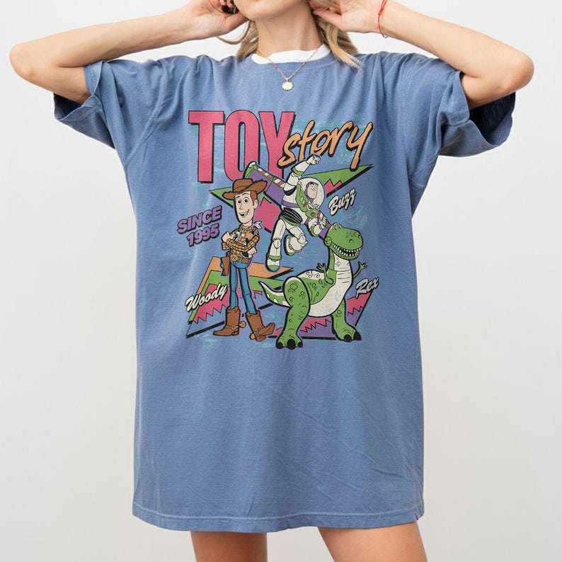 Puede incluir: Camiseta azul con un dise&ntilde;o de Toy Story que presenta a Woody, Buzz y Rex. El dise&ntilde;o incluye el texto "TOY STORY" en rosa, "SINCE 1995" y los nombres de los personajes. La camiseta tiene mangas cortas y cuello redondo.