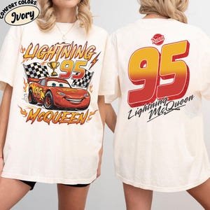 K&ouml;nnte beinhalten: Cremefarbenes T-Shirt mit einem Aufdruck von Lightning McQueen aus dem Film Cars. Das Design auf der Vorderseite zeigt "Lightning 95 McQueen" mit einem Rennwagen, Zielflaggen und einer Troph&auml;e. Auf der R&uuml;ckseite stehen "95" und "Lightning McQueen".