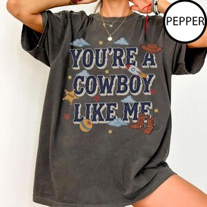 Puede incluir: Una camiseta gris carb&oacute;n de manga corta con el texto YOU'RE A COWBOY LIKE ME en letras azules. El gr&aacute;fico incluye un sombrero de vaquero marr&oacute;n, un cohete, una estrella de sheriff amarilla, una pelota colorida y botas vaqueras con nubes azules.