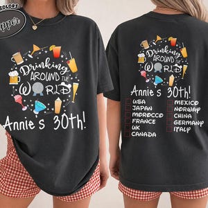 May include: A charcoal gray t-shirt featuring a graphic of various cocktails and the text Drinking Around The World and Annie's 30th. The back design includes a checklist of countries including USA, Japan, Morocco, France, UK, Canada, Mexico, Norway, China, Germany, and Italy.