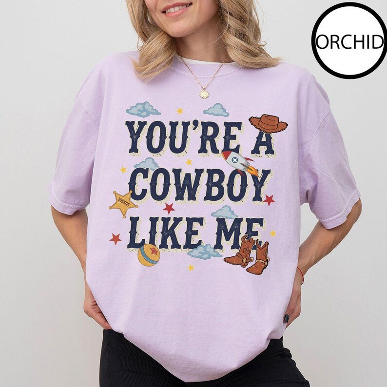 Puede incluir: Una camiseta de manga corta de color morado claro con el texto azul oscuro "YOU'RE A COWBOY LIKE ME" rodeado de ilustraciones gr&aacute;ficas de un sombrero de vaquero marr&oacute;n, un cohete, una estrella de sheriff amarilla, una pelota colorida, estrellas rojas y nubes azul claro.