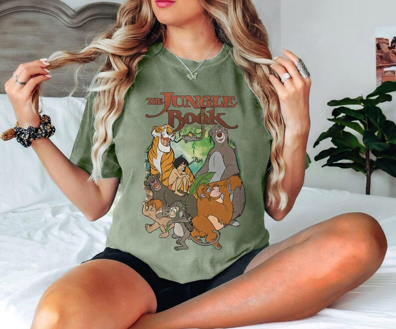 May include: Olive green t-shirt featuring a graphic from "The Jungle Book" with characters like Baloo, Bagheera, and Mowgli. The text "The Jungle Book" is in a brown, vintage-style font. The shirt is a relaxed fit.