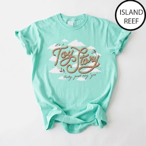 May include: A light mint green short-sleeve t-shirt featuring a graphic of white clouds and the text "Toy Story" written in a brown rope-like font, with the additional text "Baby, just say 'yes'" below it. The shirt color is labeled Island Reef.