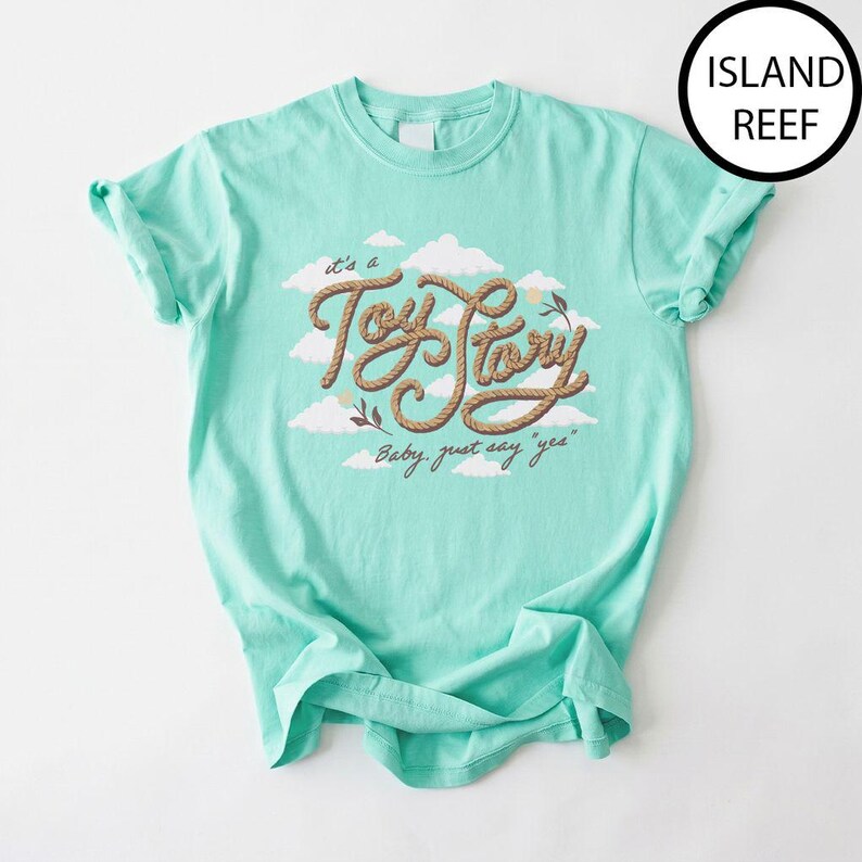 May include: A light mint green short-sleeve t-shirt featuring a graphic of white clouds and the text "Toy Story" written in a brown rope-like font, with the additional text "Baby, just say 'yes'" below it. The shirt color is labeled Island Reef.