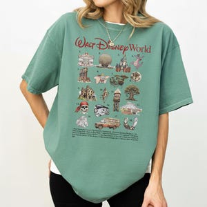 Puede incluir: Una camiseta verde de manga corta con las palabras "Walt Disney World" en escritura roja. La camiseta presenta un gr&aacute;fico con 18 ilustraciones de atracciones de Disney World, incluyendo un castillo, Spaceship Earth y una calavera pirata.