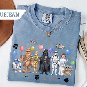 May include: Bluejean Comfort Colors t-shirt featuring Star Wars characters with balloons. The design includes R2-D2, Chewbacca, C-3PO, Darth Vader, a Stormtrooper, Princess Leia, and Baby Yoda. A gold necklace is draped across the shirt.