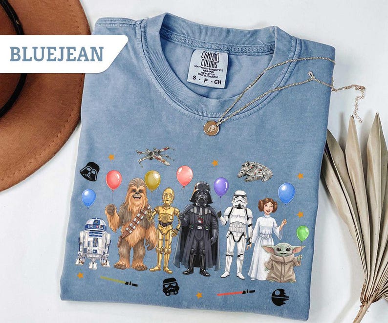 May include: Bluejean Comfort Colors t-shirt featuring Star Wars characters with balloons. The design includes R2-D2, Chewbacca, C-3PO, Darth Vader, a Stormtrooper, Princess Leia, and Baby Yoda. A gold necklace is draped across the shirt.