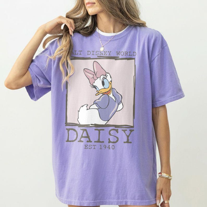 Puede incluir: Camiseta lila con un dise&ntilde;o de Daisy Duck de Disney. El dise&ntilde;o incluye el texto "DAISY EST 1940" y "Walt Disney World" en un marco rectangular. La camiseta tiene mangas cortas.
