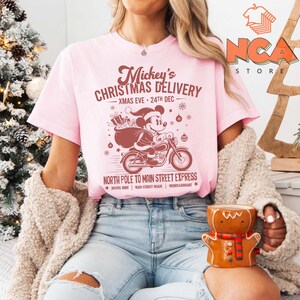 May include: A pale pink t-shirt with a festive graphic of Mickey Mouse on a motorbike, carrying gifts. Text includes "Mickey's Christmas Delivery" and "North Pole to Main Street Express". A seasonal design for Christmas.