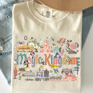 Op de afbeelding: Cr&egrave;mekleurig T-shirt met de woorden "Magic Kingdom" in kleurrijke letters, met stripfiguren en parkafbeeldingen. Het shirt bevat afbeeldingen van Mickey Mouse, Minnie Mouse, Donald Duck en andere personages.