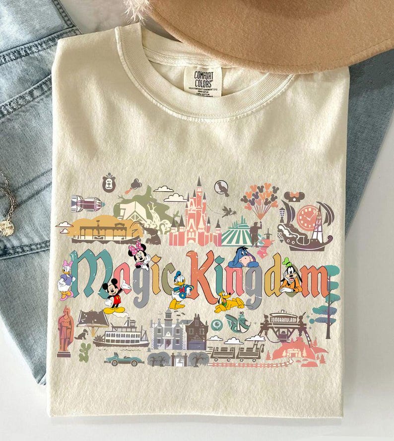 Op de afbeelding: Cr&egrave;mekleurig T-shirt met de woorden "Magic Kingdom" in kleurrijke letters, met stripfiguren en parkafbeeldingen. Het shirt bevat afbeeldingen van Mickey Mouse, Minnie Mouse, Donald Duck en andere personages.