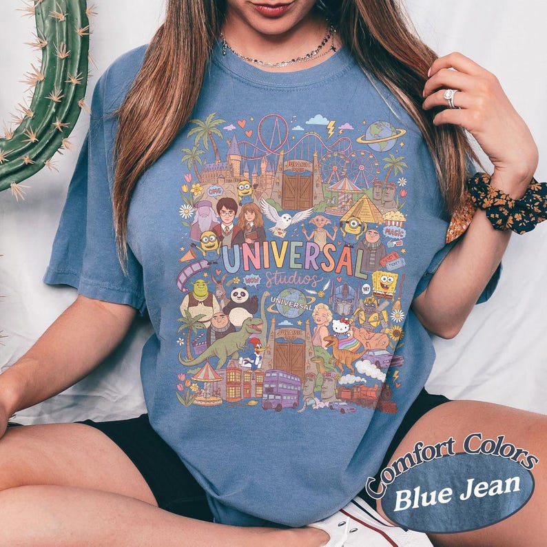 Może przedstawiać: Niebieska koszulka z kr&oacute;tkim rękawem z kolorowym nadrukiem graficznym przedstawiającym postacie i punkty orientacyjne Universal Studios. Wz&oacute;r zawiera ilustracje r&oacute;żnych animowanych postaci, kolejkę g&oacute;rską oraz napis Universal Studios.