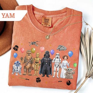 May include: Orange Comfort Colors t-shirt featuring Star Wars characters and balloons. The design includes Darth Vader, Chewbacca, C-3PO, R2-D2, a Stormtrooper, Princess Leia, and Baby Yoda. A gold necklace is draped across the shirt.