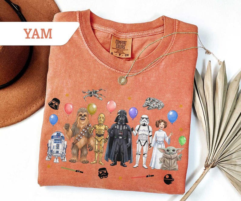 May include: Orange Comfort Colors t-shirt featuring Star Wars characters and balloons. The design includes Darth Vader, Chewbacca, C-3PO, R2-D2, a Stormtrooper, Princess Leia, and Baby Yoda. A gold necklace is draped across the shirt.