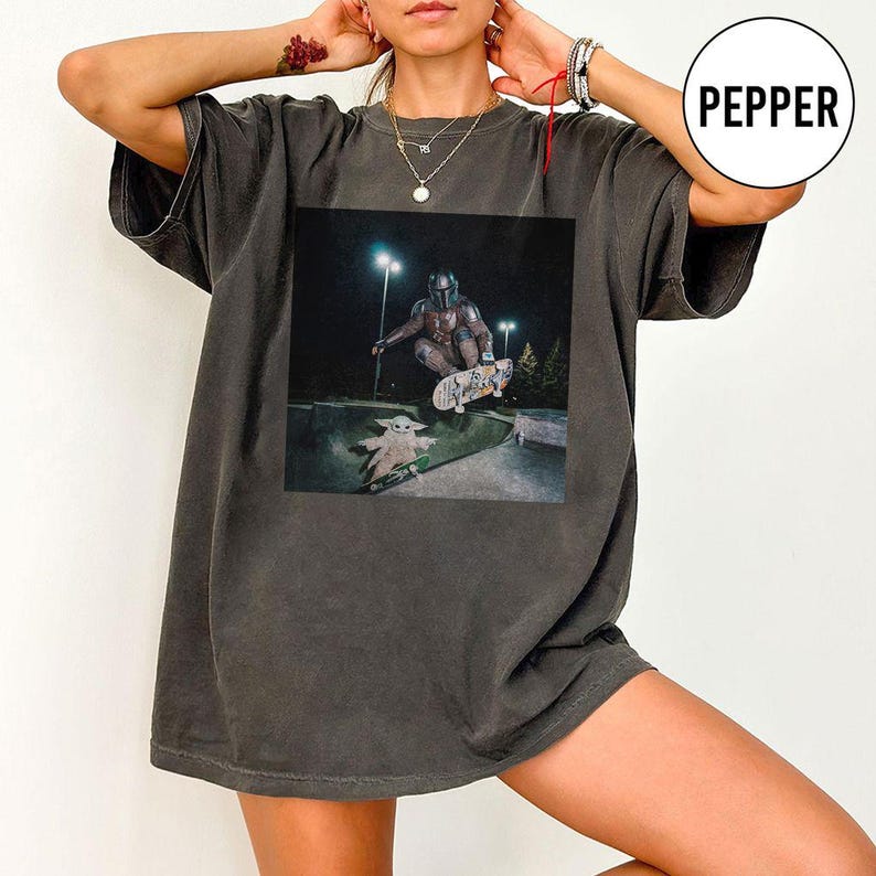May include: A charcoal gray short-sleeve t-shirt featuring a graphic print of a character in armor riding a skateboard in a skatepark at night, with a small creature on a separate skateboard below. The shirt has a crew neckline and loose fit.
