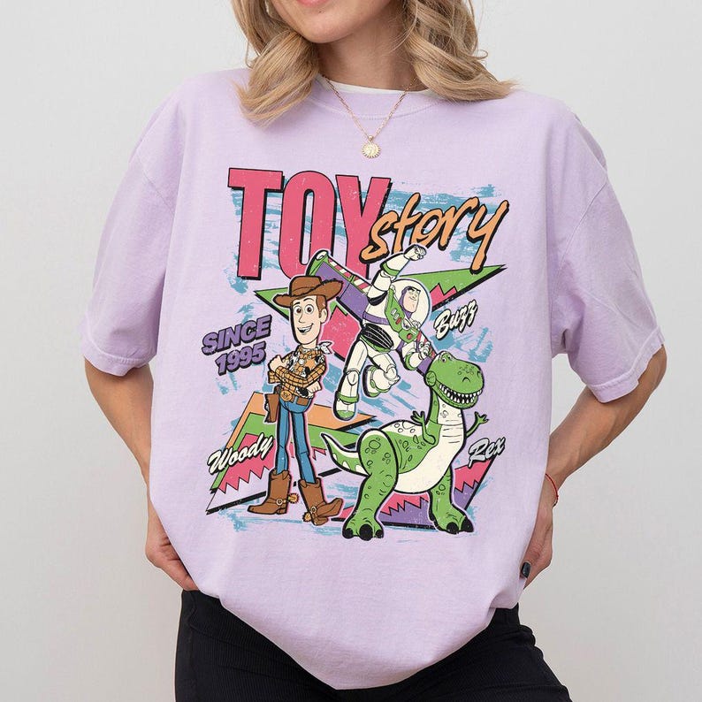 Puede incluir: Camiseta lila claro con un dise&ntilde;o de Toy Story. El dise&ntilde;o presenta a Woody, Buzz Lightyear y Rex, as&iacute; como las palabras "Toy Story" y "Since 1995" en un estilo retro.