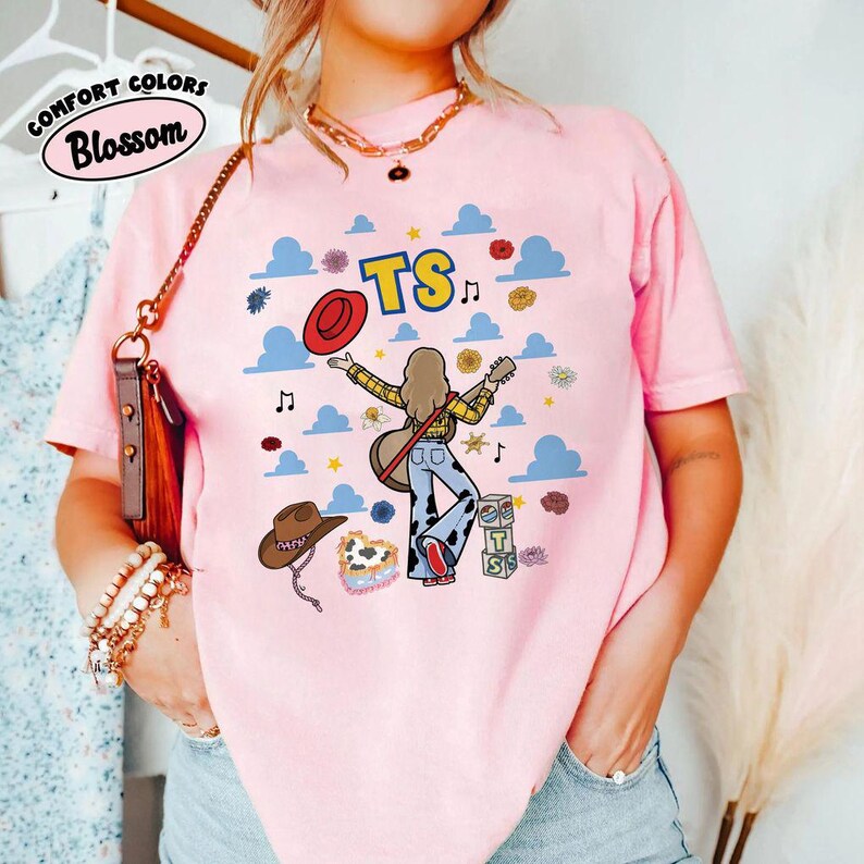May include: A light pink short-sleeve t-shirt featuring a graphic print of a person with long hair playing a guitar, wearing cow-print pants. The design includes the text TS, blue clouds, musical notes, a red hat, and a brown cowboy hat.