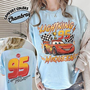 K&ouml;nnte beinhalten: Hellblaues T-Shirt mit Lightning McQueen aus dem Film Cars. Auf der Vorderseite sind die Nummer 95, eine Troph&auml;e und Zielflaggen abgebildet. Auf der R&uuml;ckseite stehen ebenfalls die Nummer 95 und "Lightning McQueen". Das Shirt ist mit "Comfort Colors Chambray" gekennzeichnet.