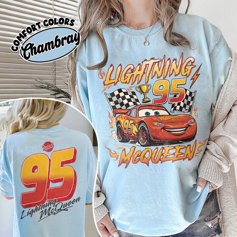 K&ouml;nnte beinhalten: Hellblaues T-Shirt mit Lightning McQueen aus dem Film Cars. Auf der Vorderseite sind die Nummer 95, eine Troph&auml;e und Zielflaggen abgebildet. Auf der R&uuml;ckseite stehen ebenfalls die Nummer 95 und "Lightning McQueen". Das Shirt ist mit "Comfort Colors Chambray" gekennzeichnet.