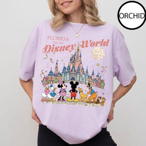 Puede incluir: Camiseta lila claro con un gr&aacute;fico colorido de Disney World en Florida. El dise&ntilde;o incluye el texto "Florida Disney World Est. 1971" y personajes de Disney como Mickey y Minnie Mouse y Donald Duck.