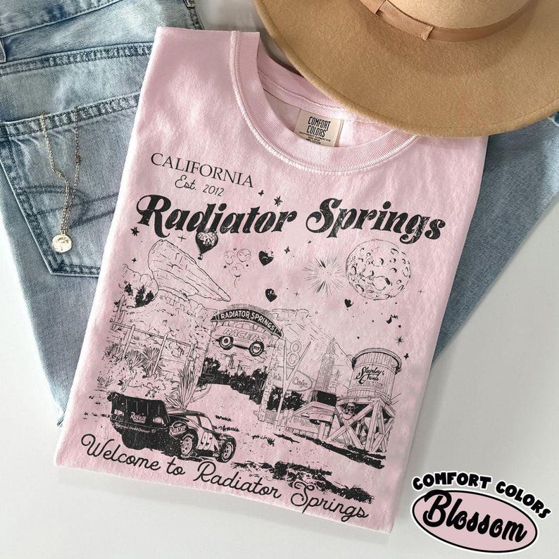 May include: A light pink t-shirt with a graphic of Radiator Springs, California, and the text "Welcome to Radiator Springs." The shirt is from Comfort Colors and is labeled "Blossom."