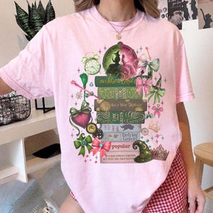 以下が含まれることがあります： ファンタジーをテーマにしたデザインの、明るいピンク色のTシャツ。 「Defying Gravity」や「Popular」などのタイトルが付いた本の山、時計、リボン、ハート、魔女の帽子などがデザインされています。「The Wizard and」の文字も確認できます。