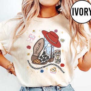 May include: An ivory t-shirt featuring a graphic print of a cowboy boot with a cow print pattern, a red hat, a stack of blocks with the letters T, S, and the number 13, a bow, flowers, and a beaded string with the text I KNEW IT WHEN YOU.